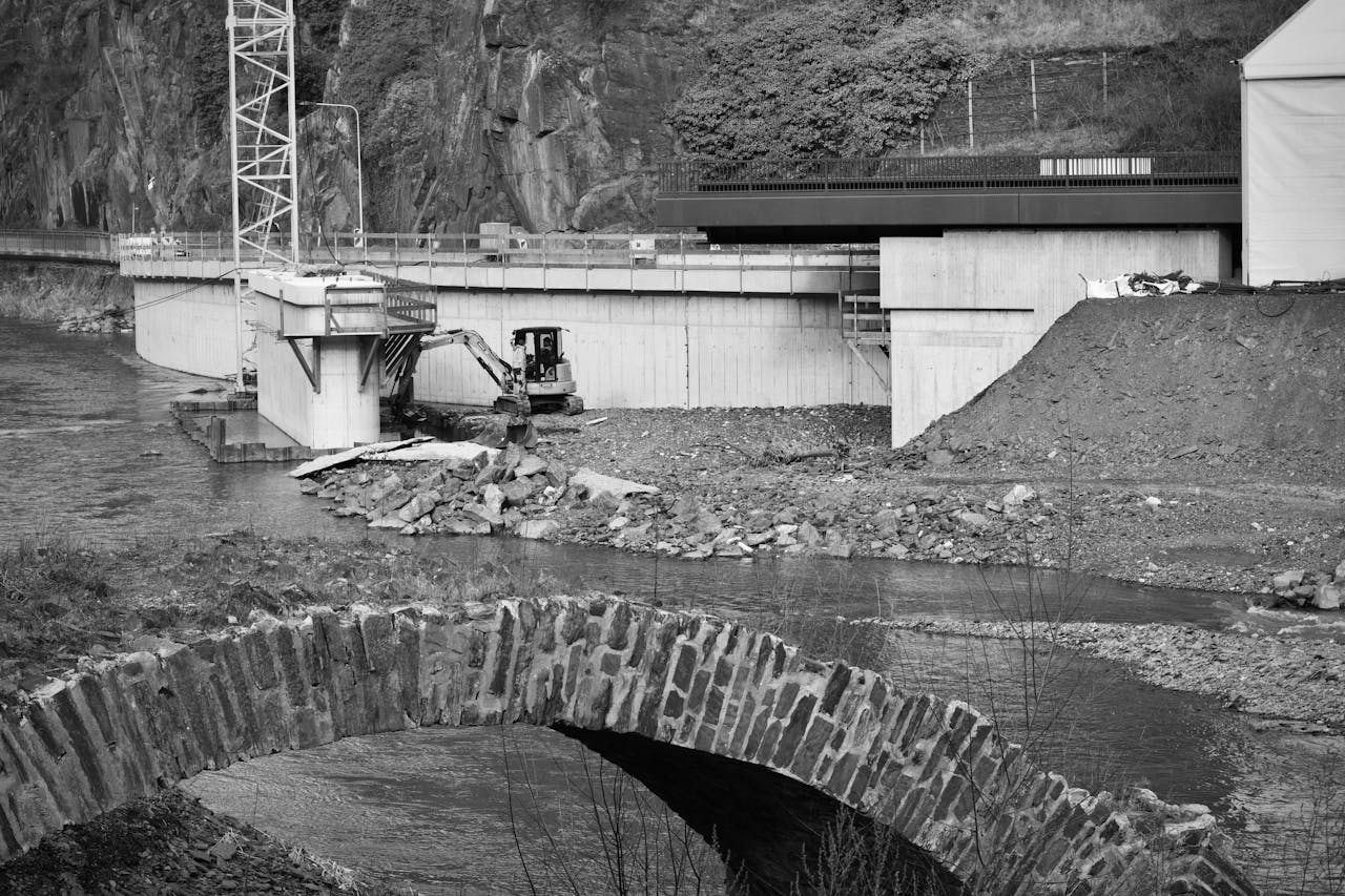 Monochrome image of bridge construction over a river with heavy machinery and stone arch.
